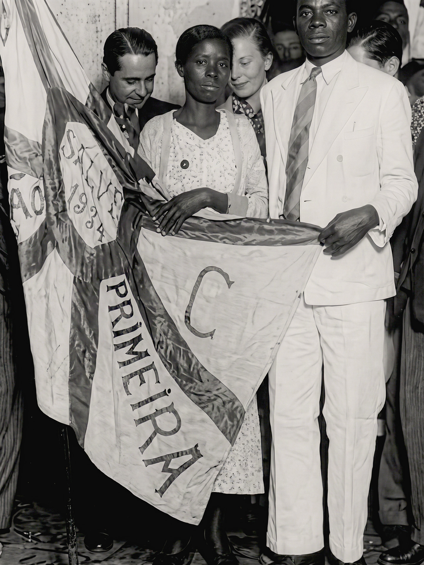Massu e Raimunda, ambos de roupa branca, segurando a bandeira do então Bloco Estação Primeira com outras pessoas ao redor durante a inauguração da Escola Municipal Humberto de Campos