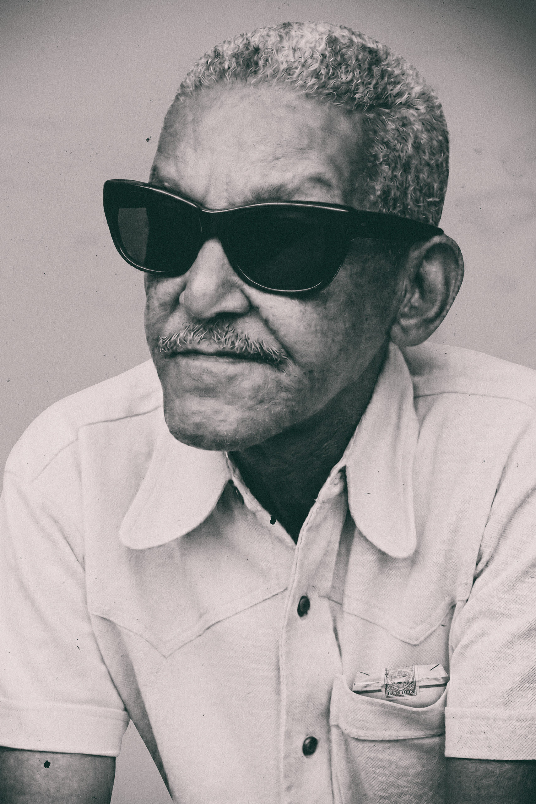 Retrato de Cartola (Angenor de Oliveira) da Mangueira usando óculos escuros e camisa clara de manga curta, olhando levemente para o lado