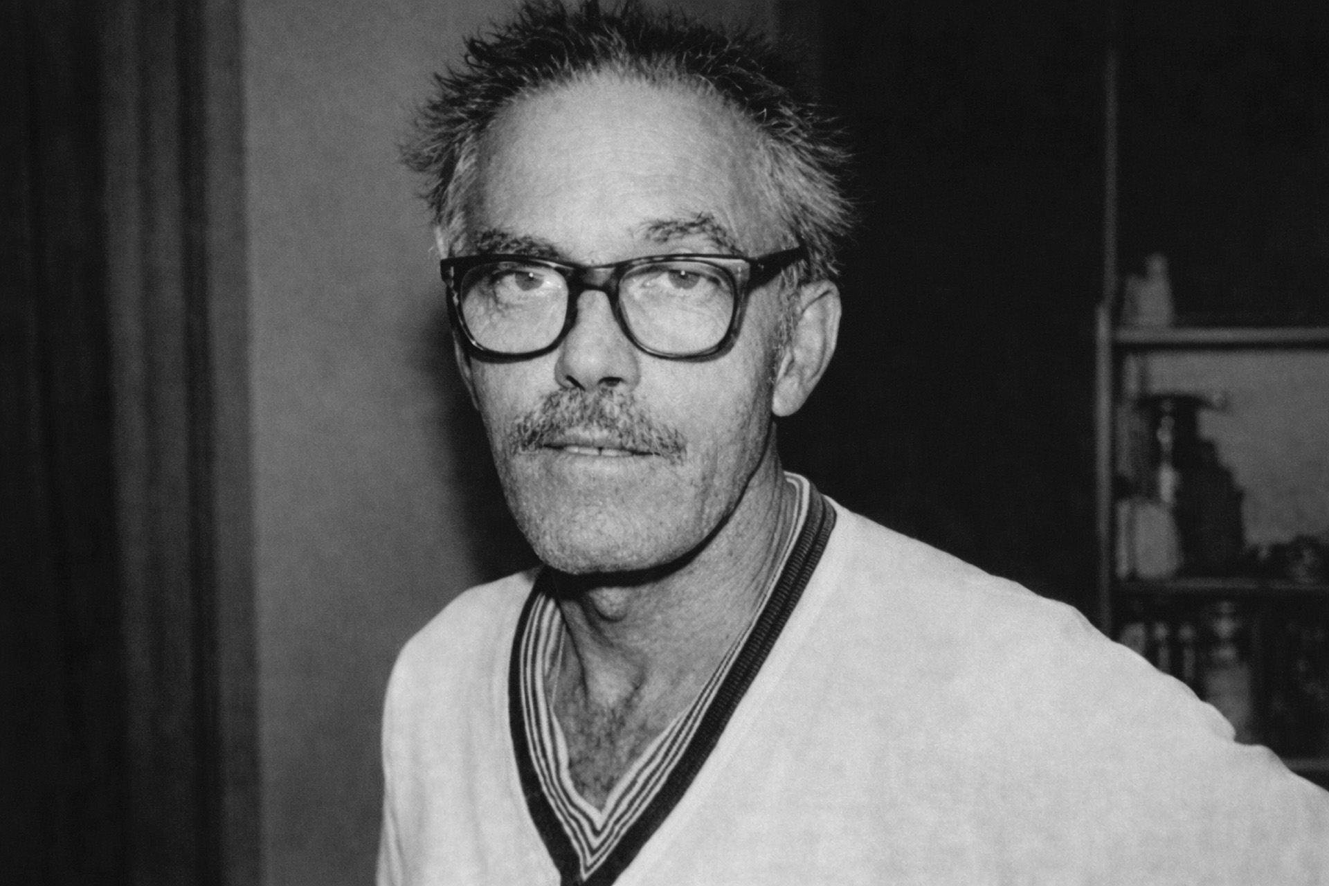 Retrato em preto e branco de Hélio Turco da Mangueira usando óculos de aros grossos, bigode e suéter de gola V com listras