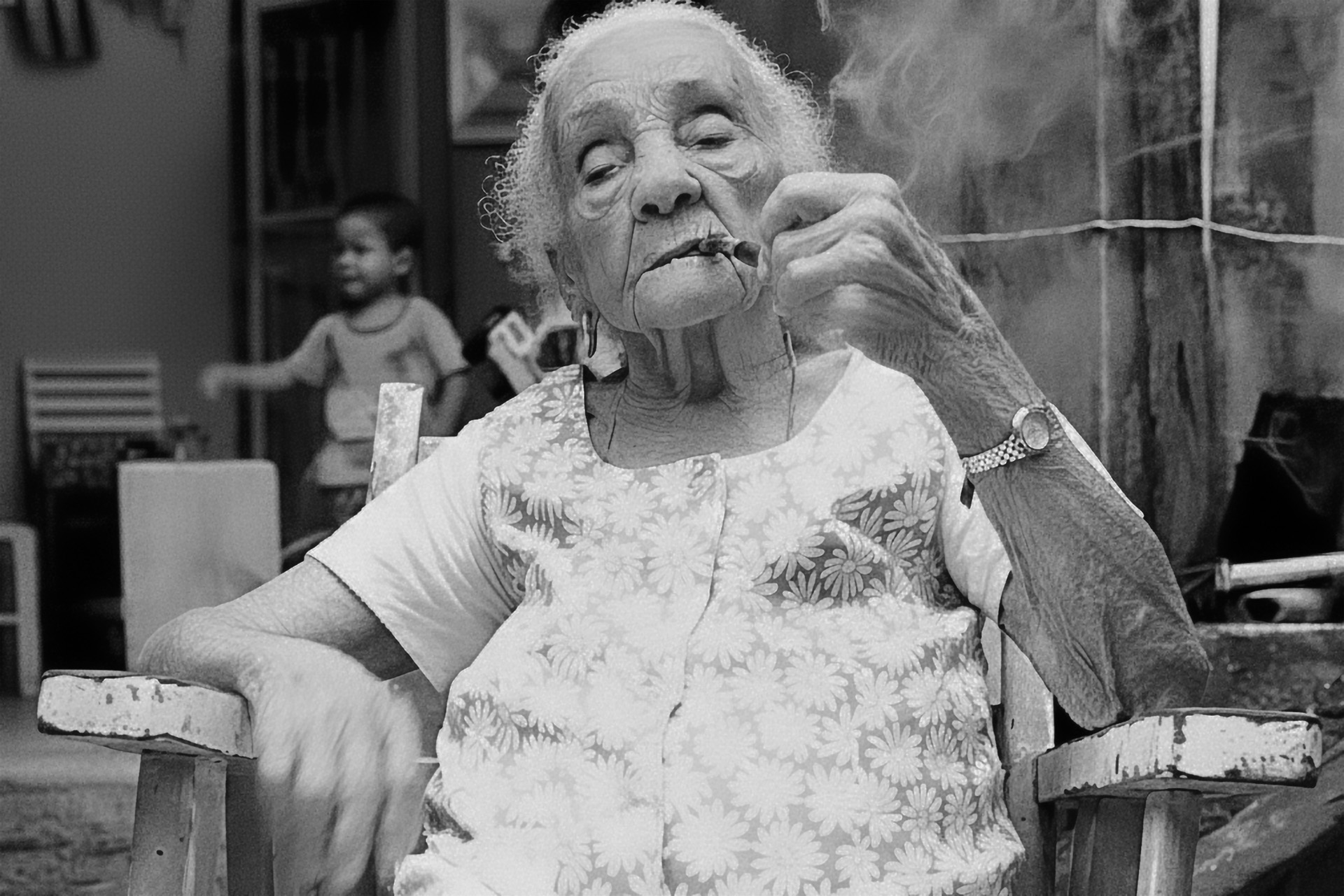 Vó Lucíola, neta de escravos e pioneira da Mangueira, sentada em uma cadeira de madeira fumando cachimbo com fumaça subindo, criança ao fundo