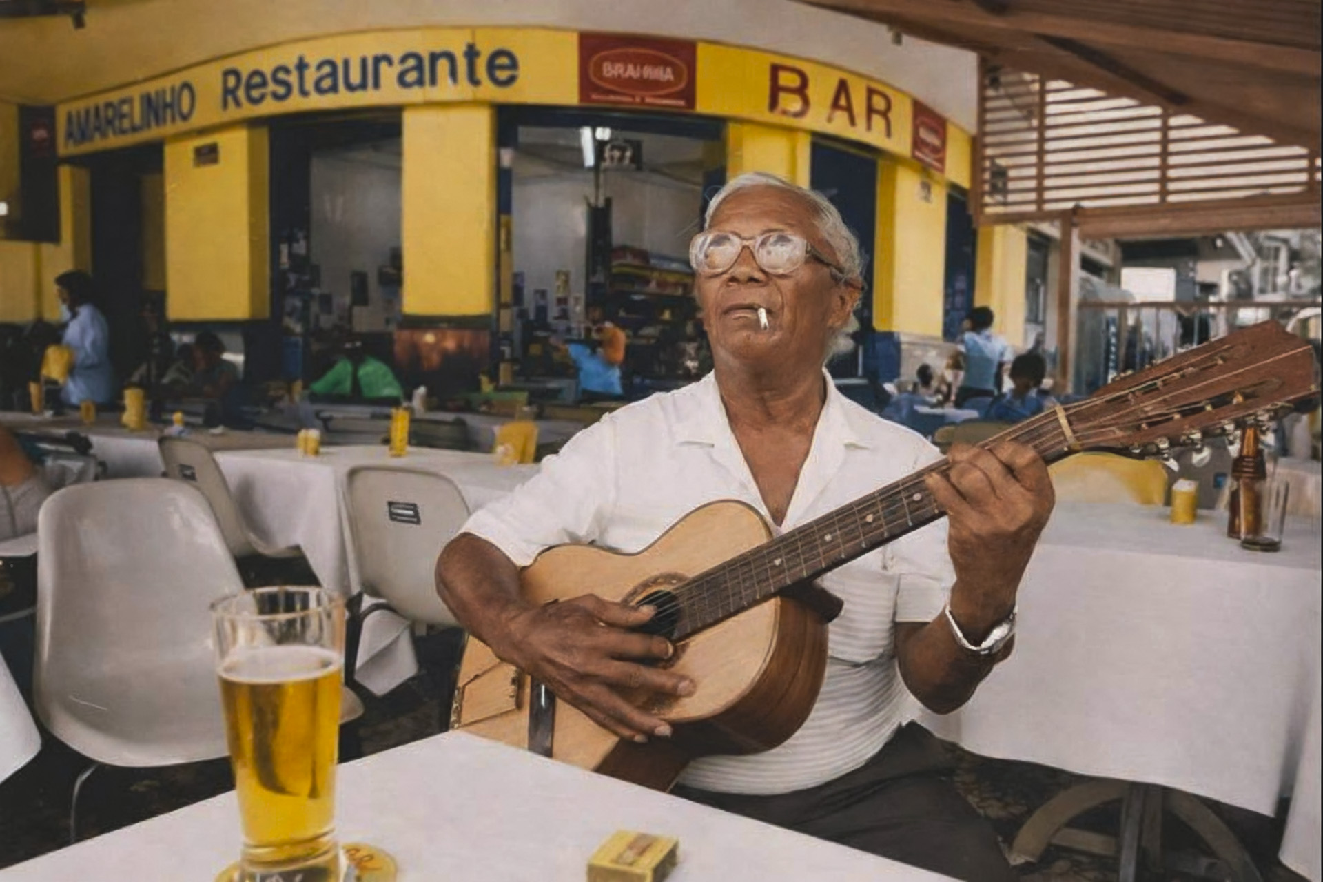 Nélson Cavaquinho sentado à mesa do restaurante Amarelinho tocando violão com um copo de cerveja à frente