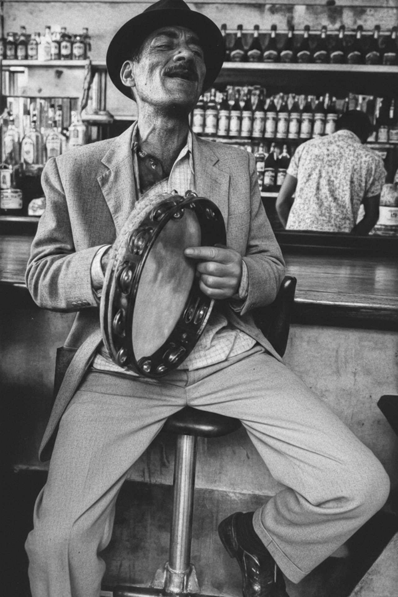 Padeirinho da Mangueira sentado em um banco de bar tocando pandeiro com prateleiras de garrafas ao fundo