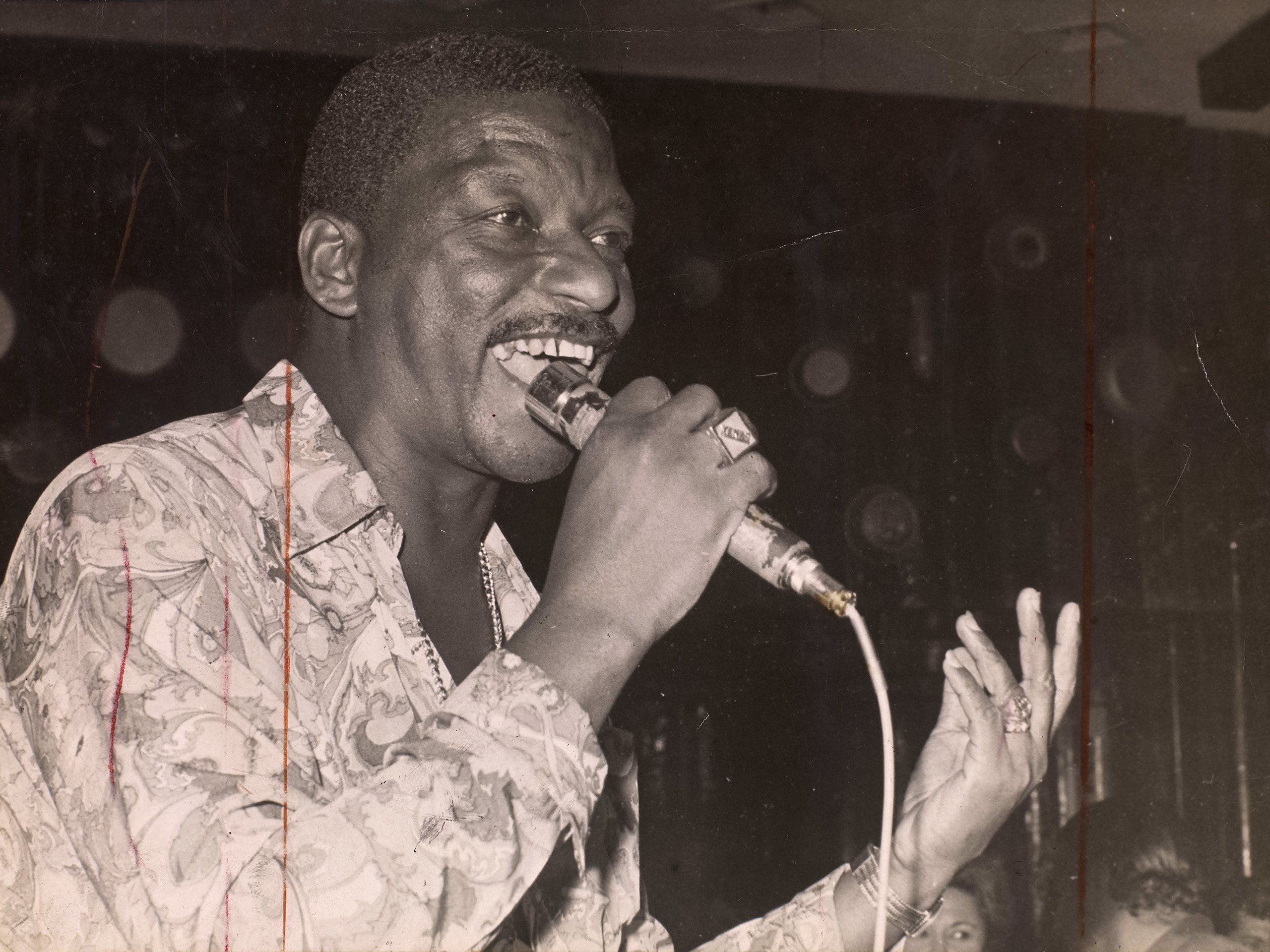 Xangô da Mangueira cantando ao microfone com camisa estampada e corrente no pescoço, mão aberta em gesto expressivo