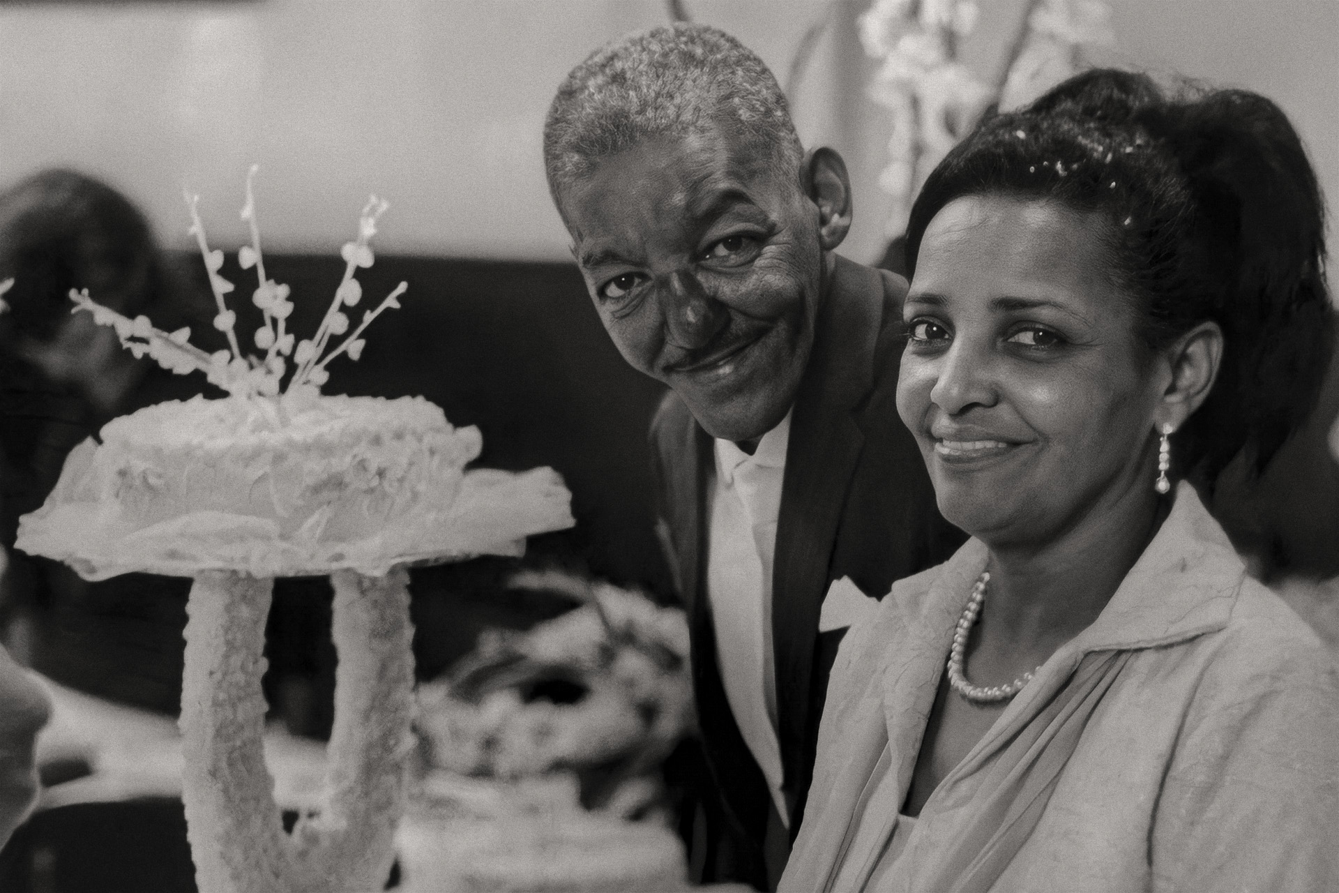 Cartola e Zica da Mangueira sorrindo ao lado do bolo de casamento, ele de smoking e ela com colar de pérolas e brincos