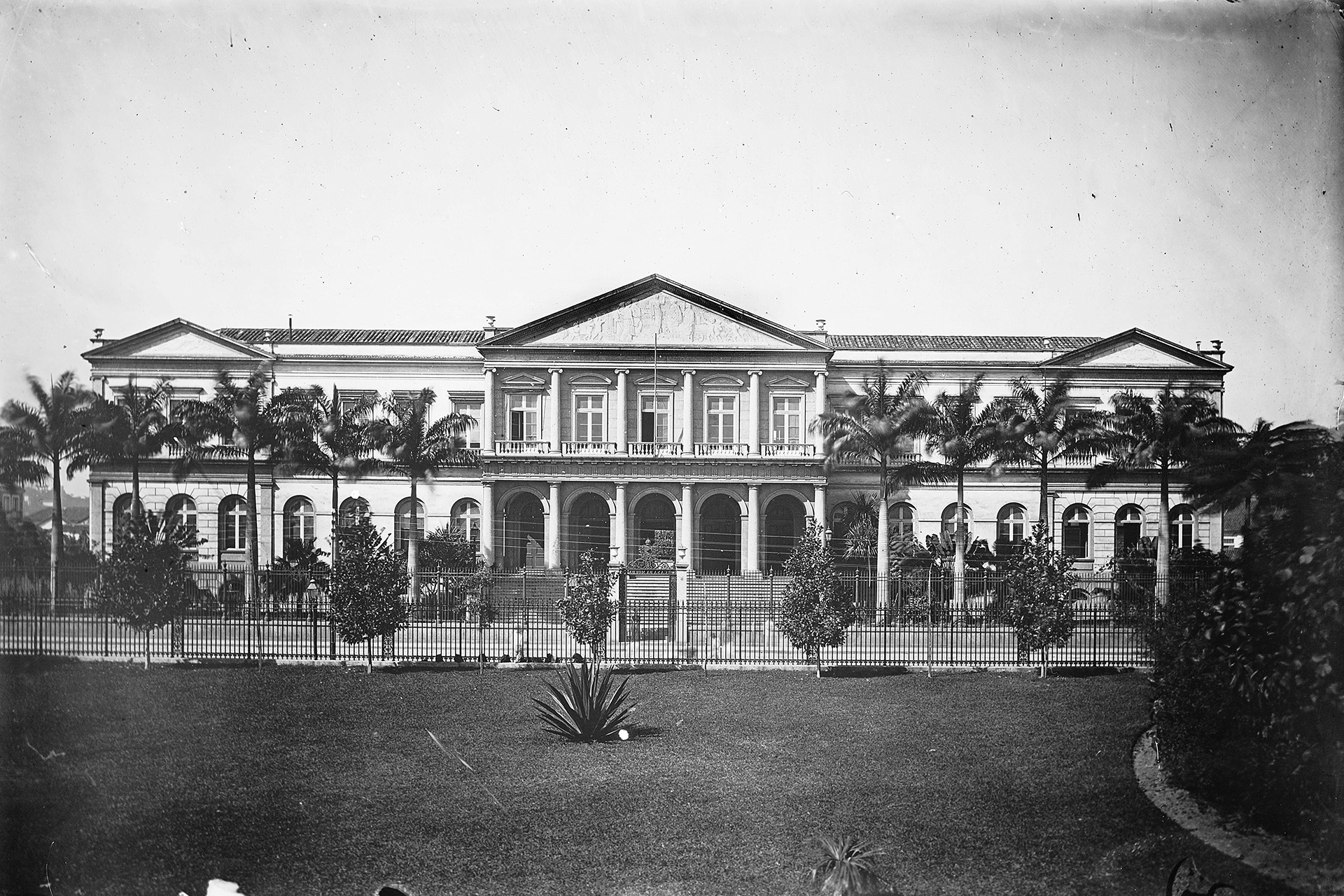 Fachada neoclássica da antiga Casa da Moeda, atual Arquivo Nacional, com jardim, palmeiras imperiais e grades de ferro em foto de 1890