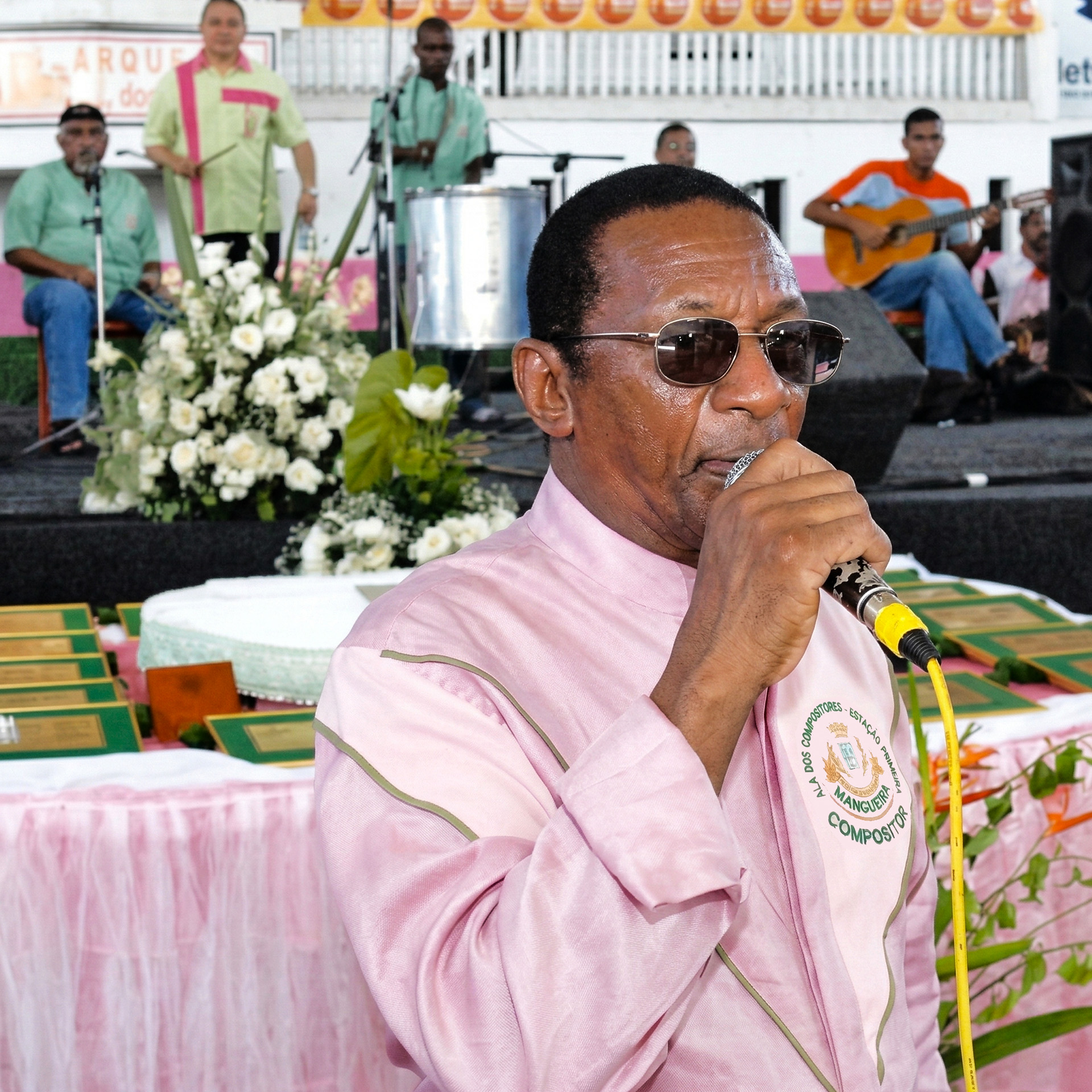 Darcy da Mangueira cantando ao microfone com camisa rosa da Ala dos Compositores e óculos escuros no Palácio do Samba, com músicos e arranjo floral ao fundo