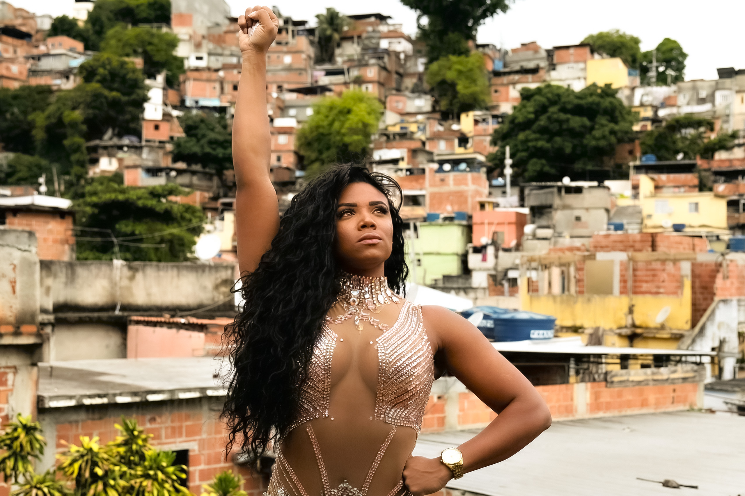 Evelyn Bastos com fantasia cravejada de cristais e punho erguido em pose altiva no morro de Mangueira, com as casas da comunidade ao fundo