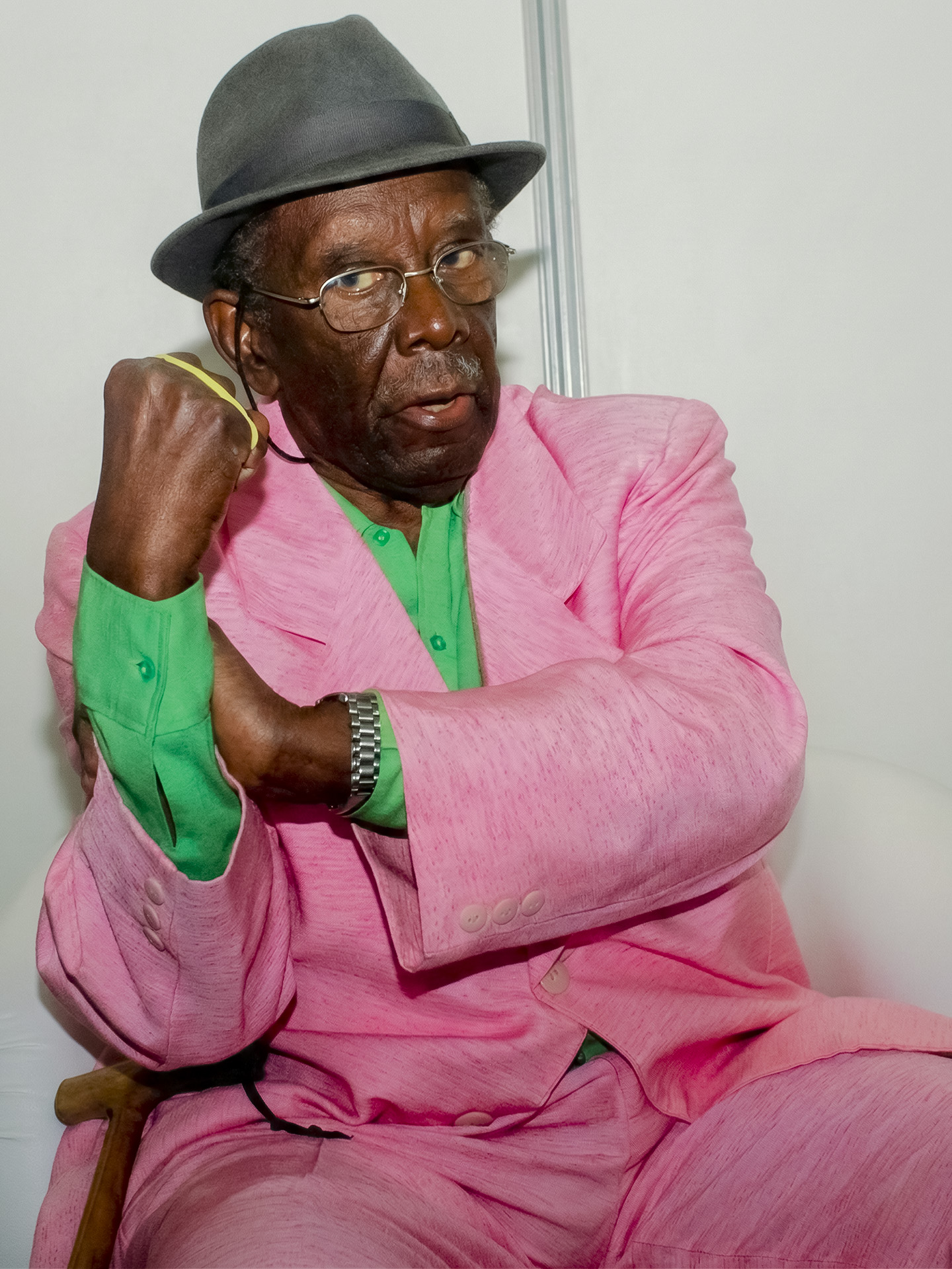 Jamelão da Mangueira sentado com terno rosa, camisa verde e chapéu preto, punho erguido em gesto expressivo