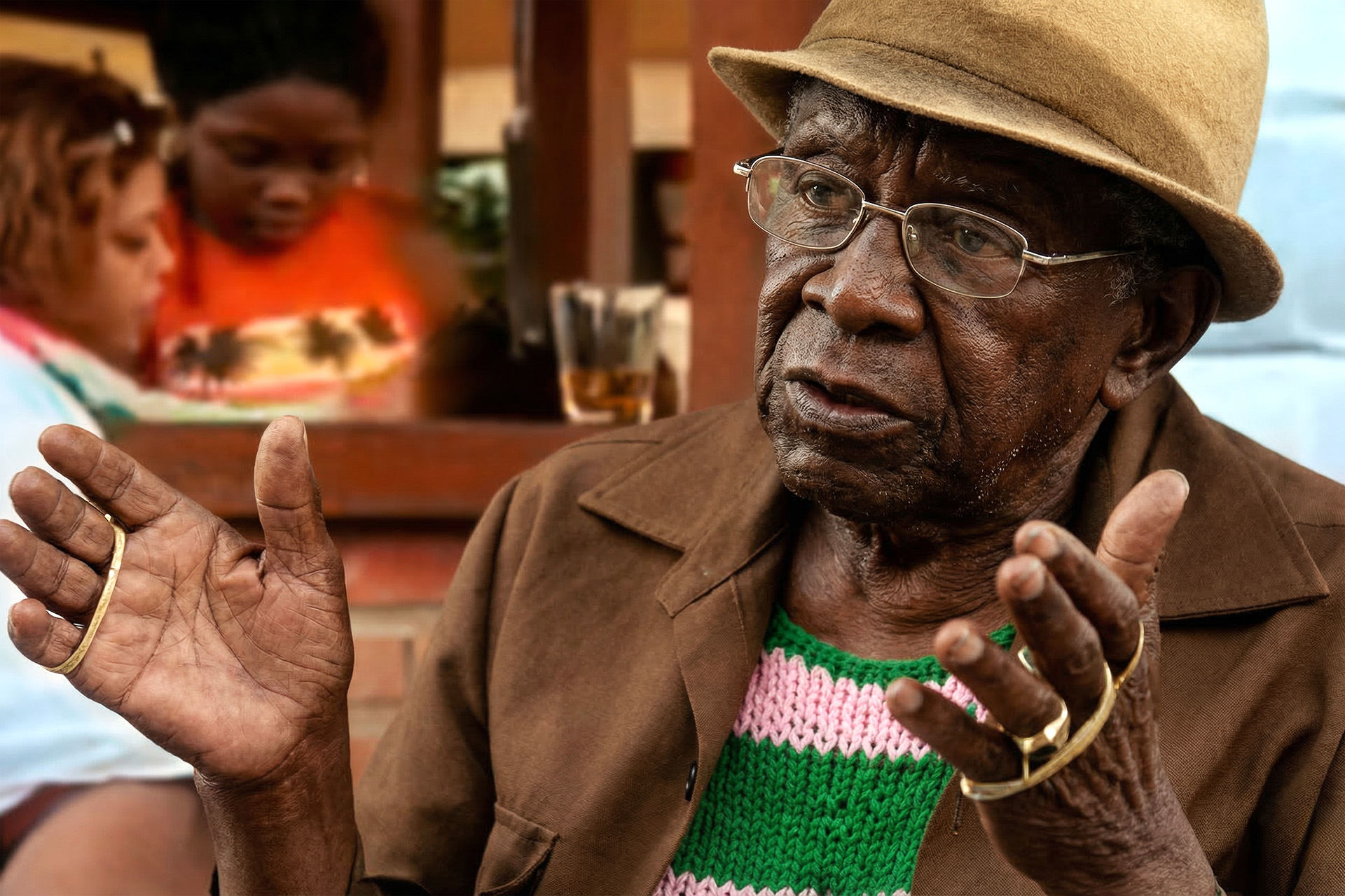 Jamelão da Mangueira de chapéu bege, óculos e suéter verde-rosa gesticulando com as duas mãos durante conversa na casa de Zeca Pagodinho em Xerém