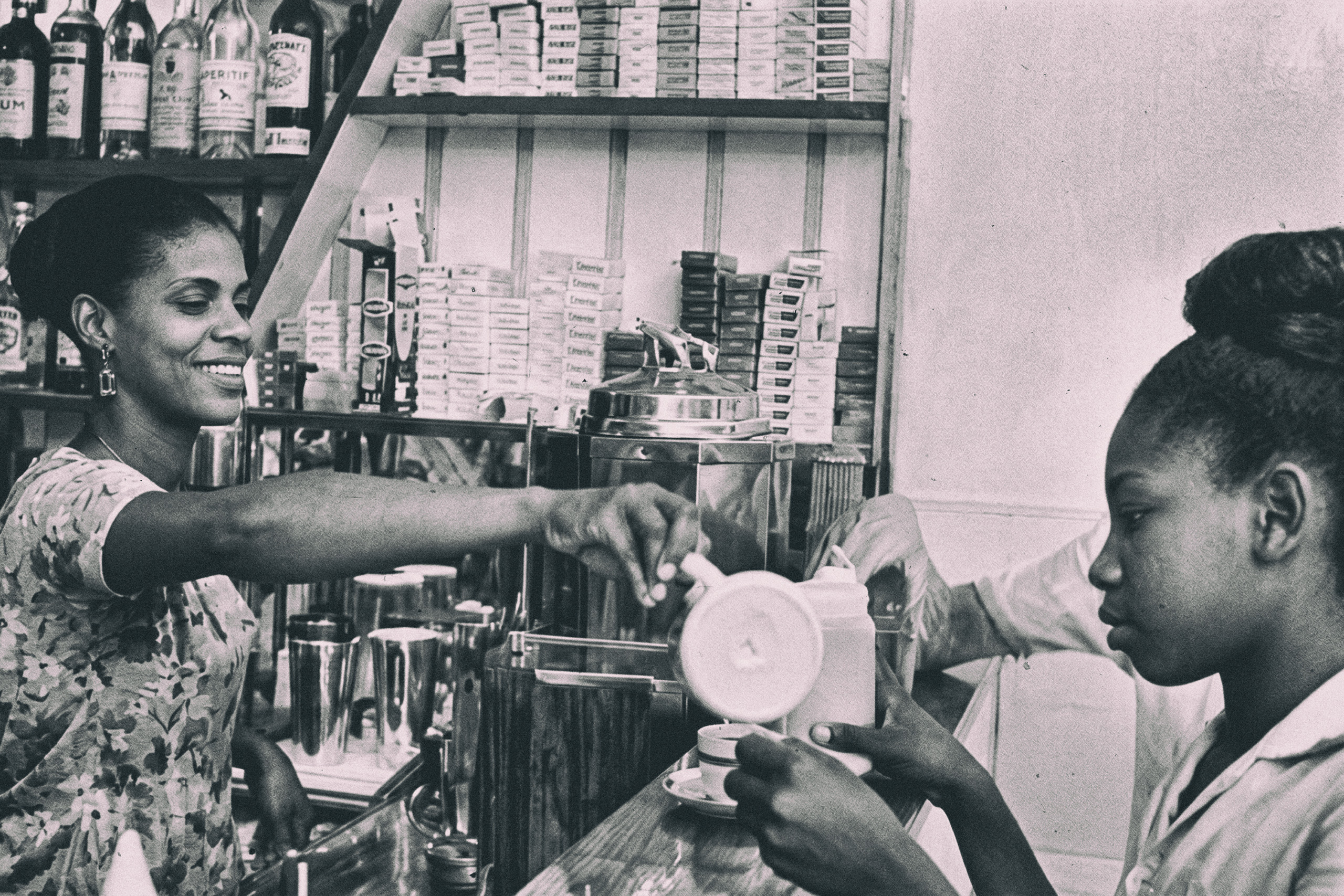 Neide da Mangueira sorrindo atrás do balcão de seu bar no CADEG, servindo café a uma cliente, com prateleiras de bebidas e caixas ao fundo