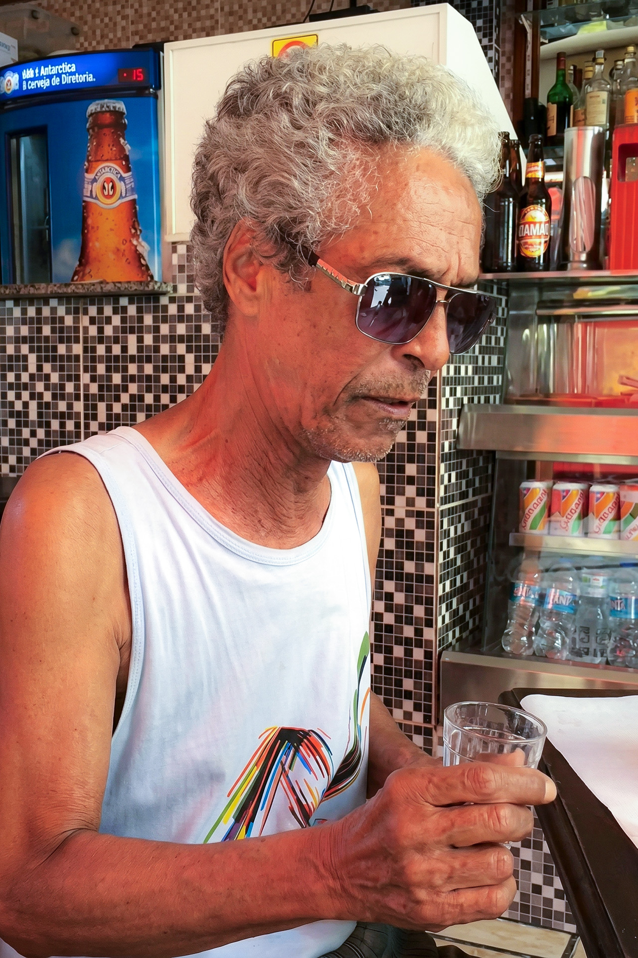 Sobrinho, intérprete da Mangueira, de óculos escuros e regata branca segurando um copo em um bar do Rio de Janeiro