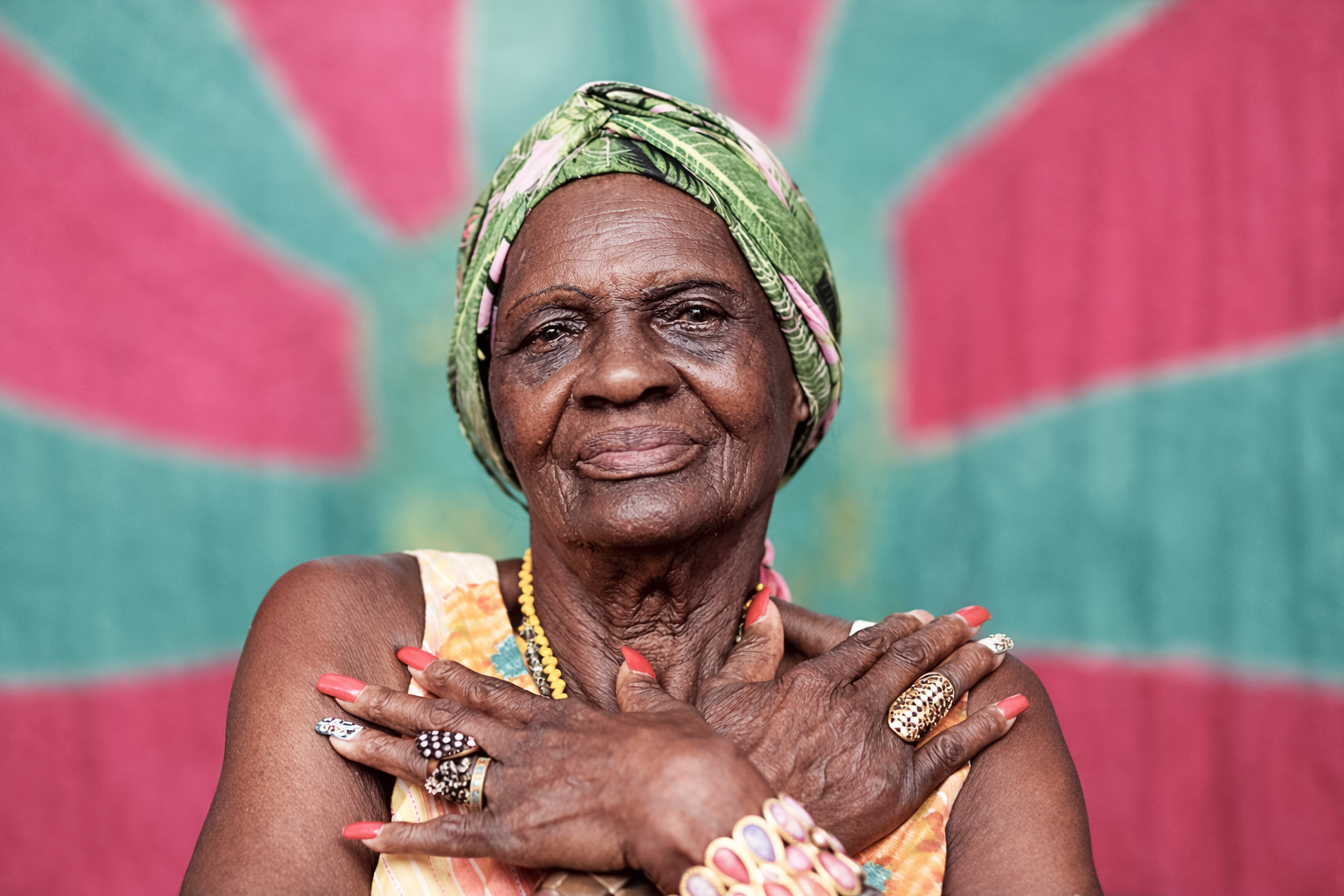 Retrato de Suluca da Mangueira com turbante verde, colares, anéis e unhas vermelhas, mãos cruzadas sobre o peito, diante de painel verde e rosa