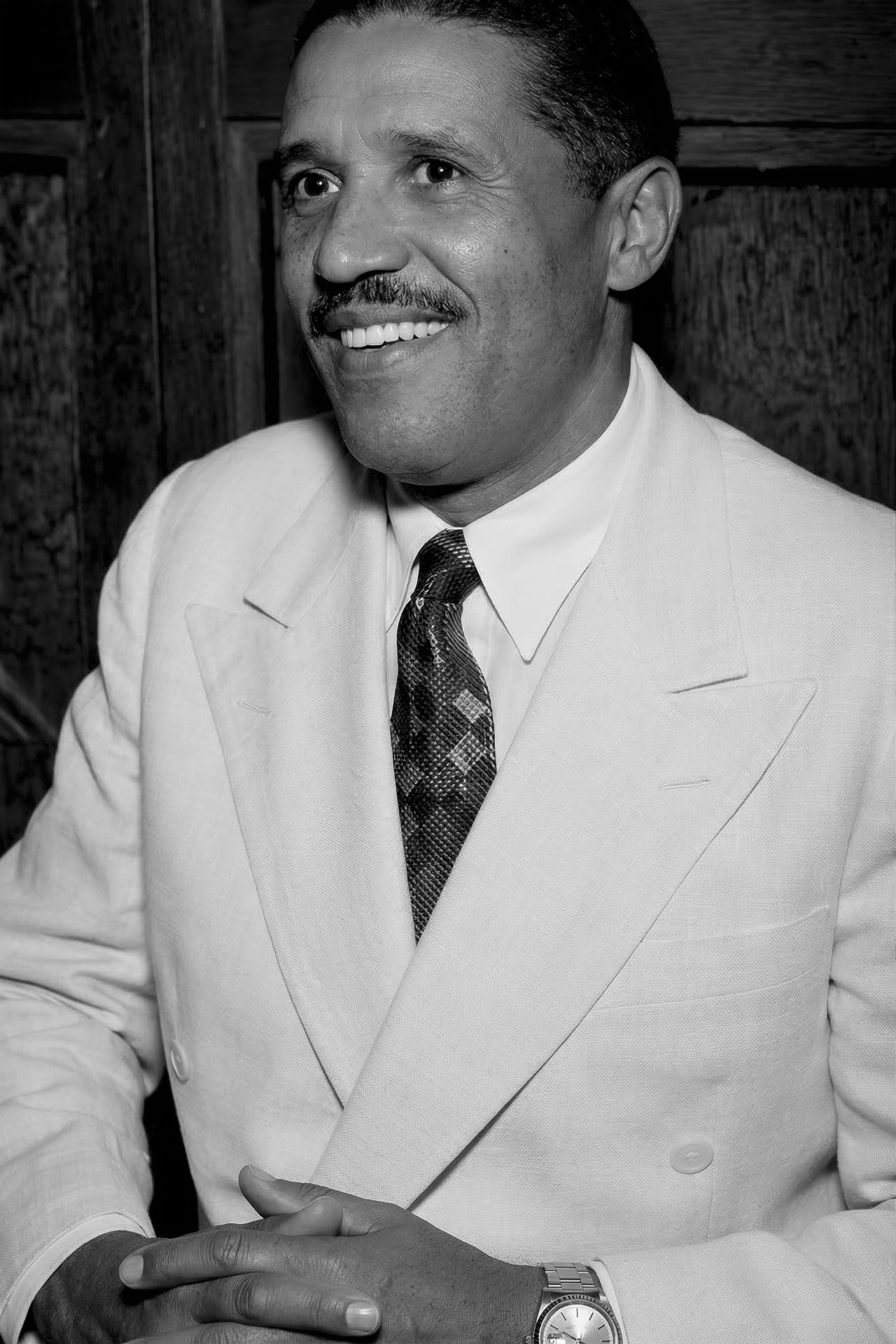 Retrato de Zé-com-Fome da Mangueira sorrindo com terno branco de lapela dupla, gravata escura e relógio de pulso, mãos entrelaçadas