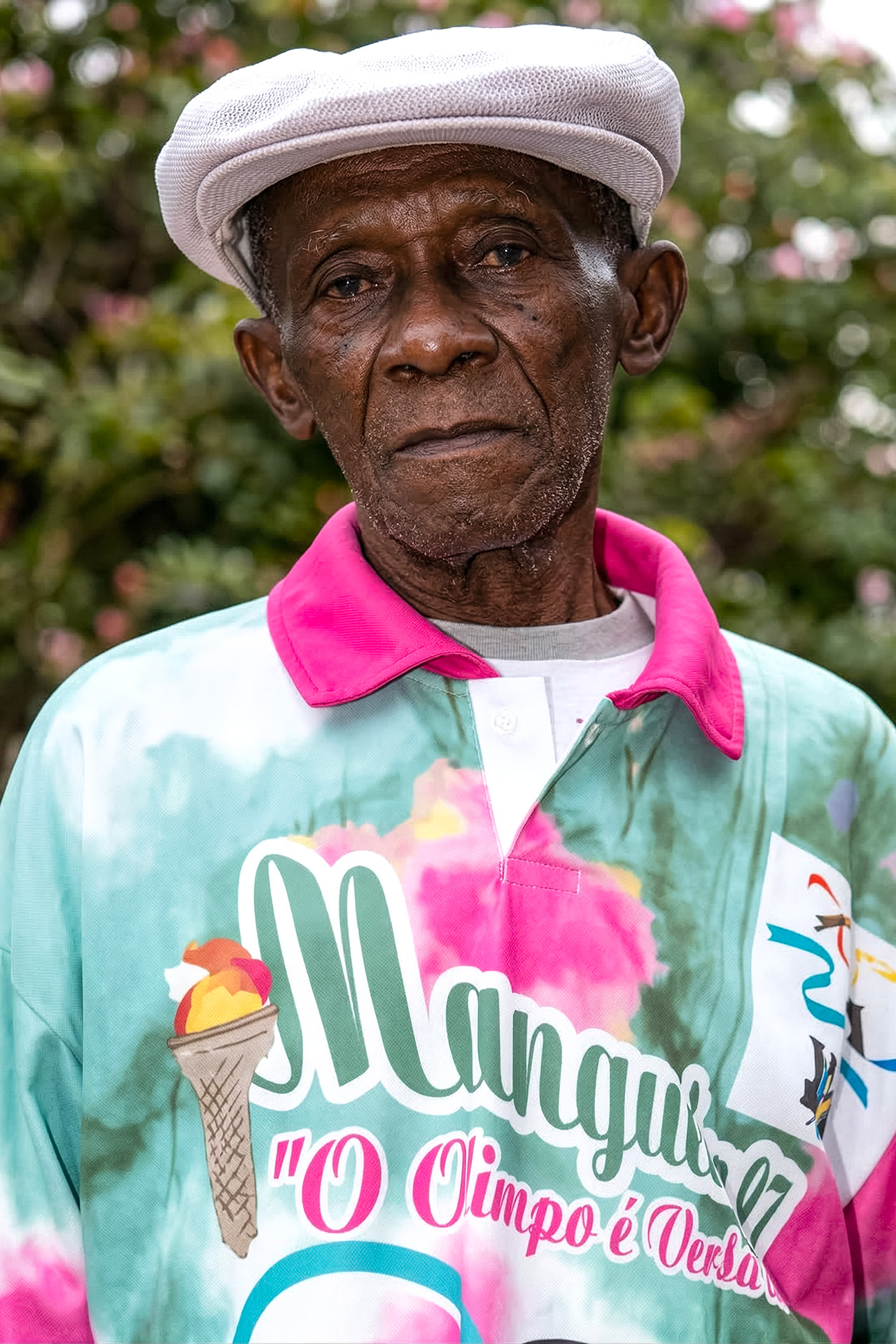 Retrato de Zé Crioulinho com boné branco e camisa da Mangueira "O Olimpo é Verde Rosa", olhar sério e vegetação ao fundo